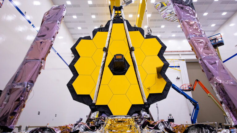 The James Webb Space Telescope with its golden hexagonal mirrors is shown inside a clean room facility, surrounded by technical equipment and scaffolding.