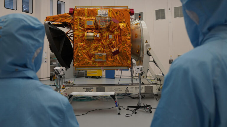 Two people in blue protective suits observe a satellite covered in reflective material, mounted on a platform inside a clean room.