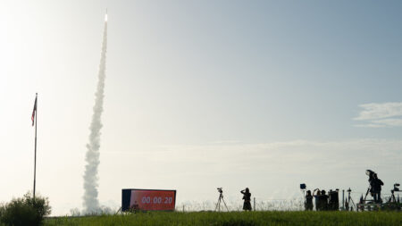 A rocket ascends into the sky, leaving a trail of smoke behind. People and cameras are gathered on the ground to the right, with an American flag and a digital countdown clock in the foreground.
