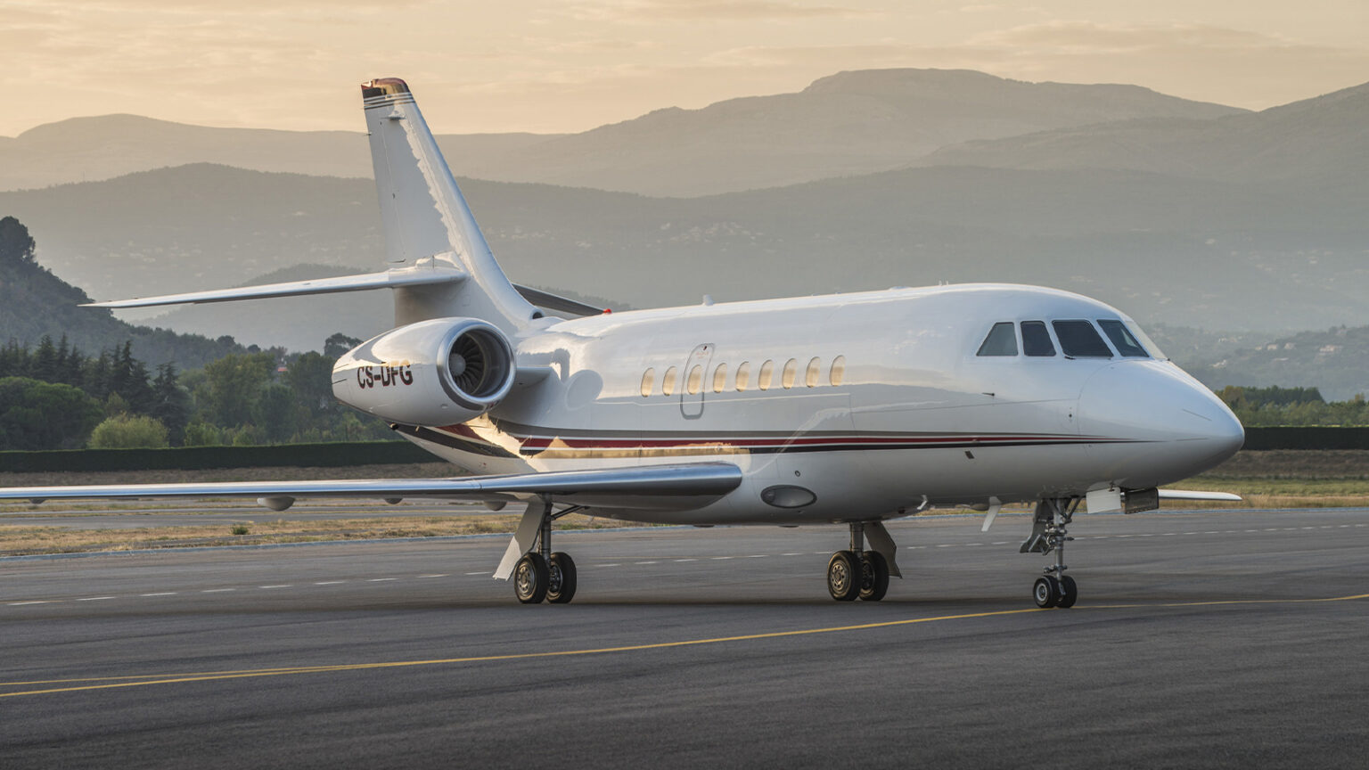 A white private jet is parked on an airstrip with mountains in the background at sunset.