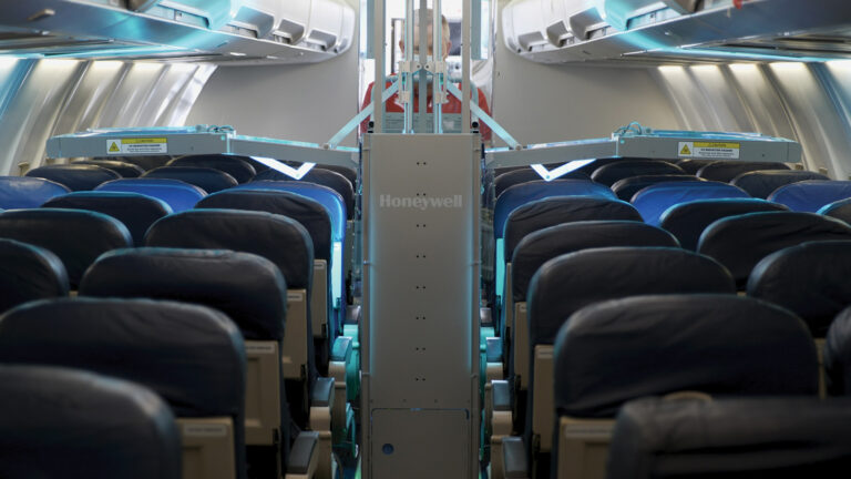 An empty aircraft cabin with rows of blue seats, overhead compartments, and a Honeywell air purification system installed in the center aisle.