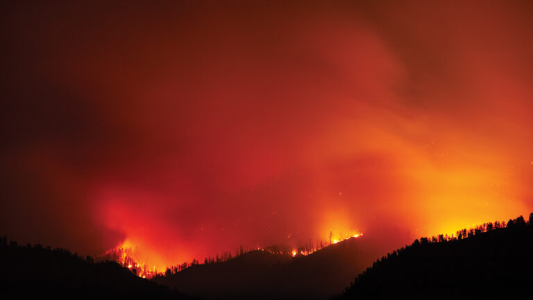 A vast wildfire blazes across a forested mountain range under a dark sky, illuminating the landscape with intense orange and red hues.