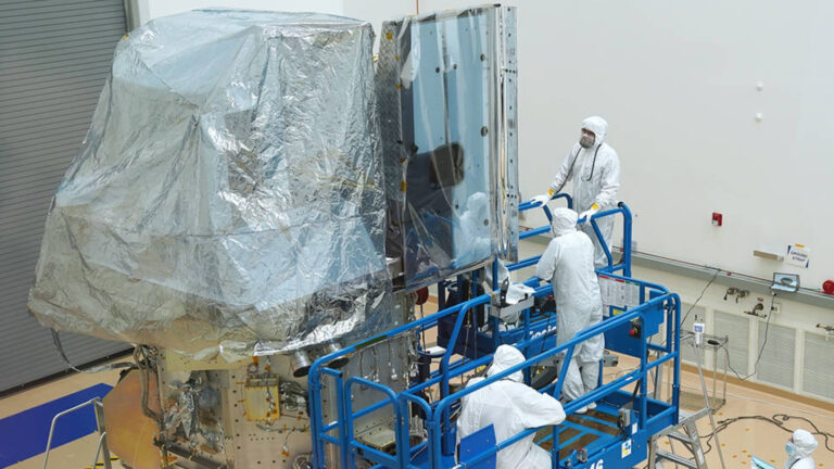 Three technicians in white suits work on a spacecraft in a clean room, surrounded by equipment, with part of the spacecraft covered in protective foil.