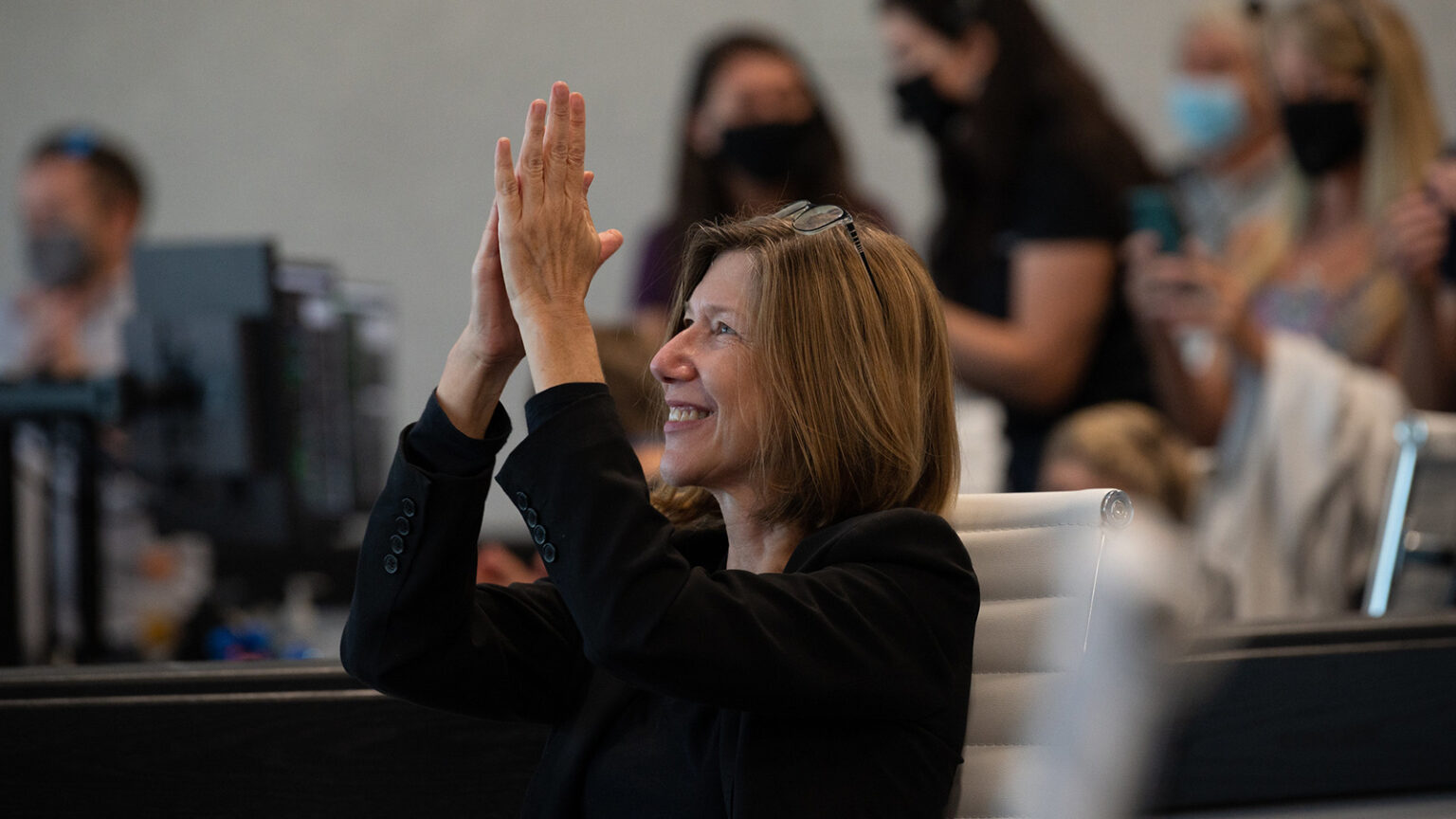 A woman, smiling and wearing a black blazer, applauds while seated in an office setting. Other people, some with masks, are visible in the background.