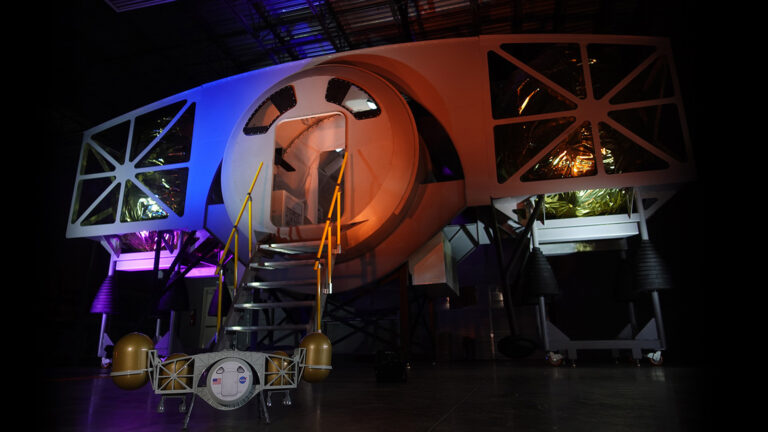A futuristic spacecraft model with illuminated panels and a central circular entrance, flanked by staircases, bathed in blue and purple lighting. A small research instrument is placed in the foreground.