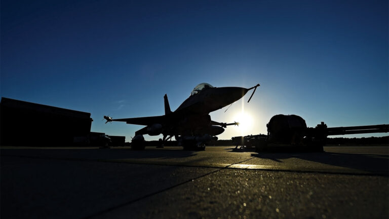 Silhouette of a fighter jet on the ground at sunset, with the sun creating a dramatic backlighting effect. The surrounding area features aircraft equipment and minimal structures.
