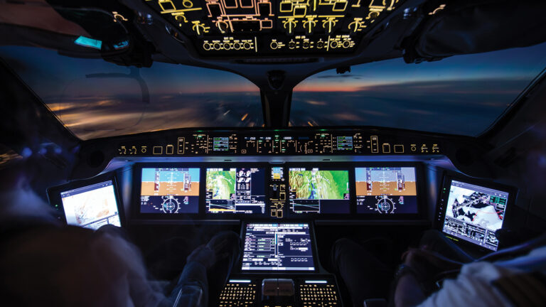 View from inside a modern airplane cockpit, displaying multiple digital screens and controls, with a blurred view of the sunset horizon through the windscreen.