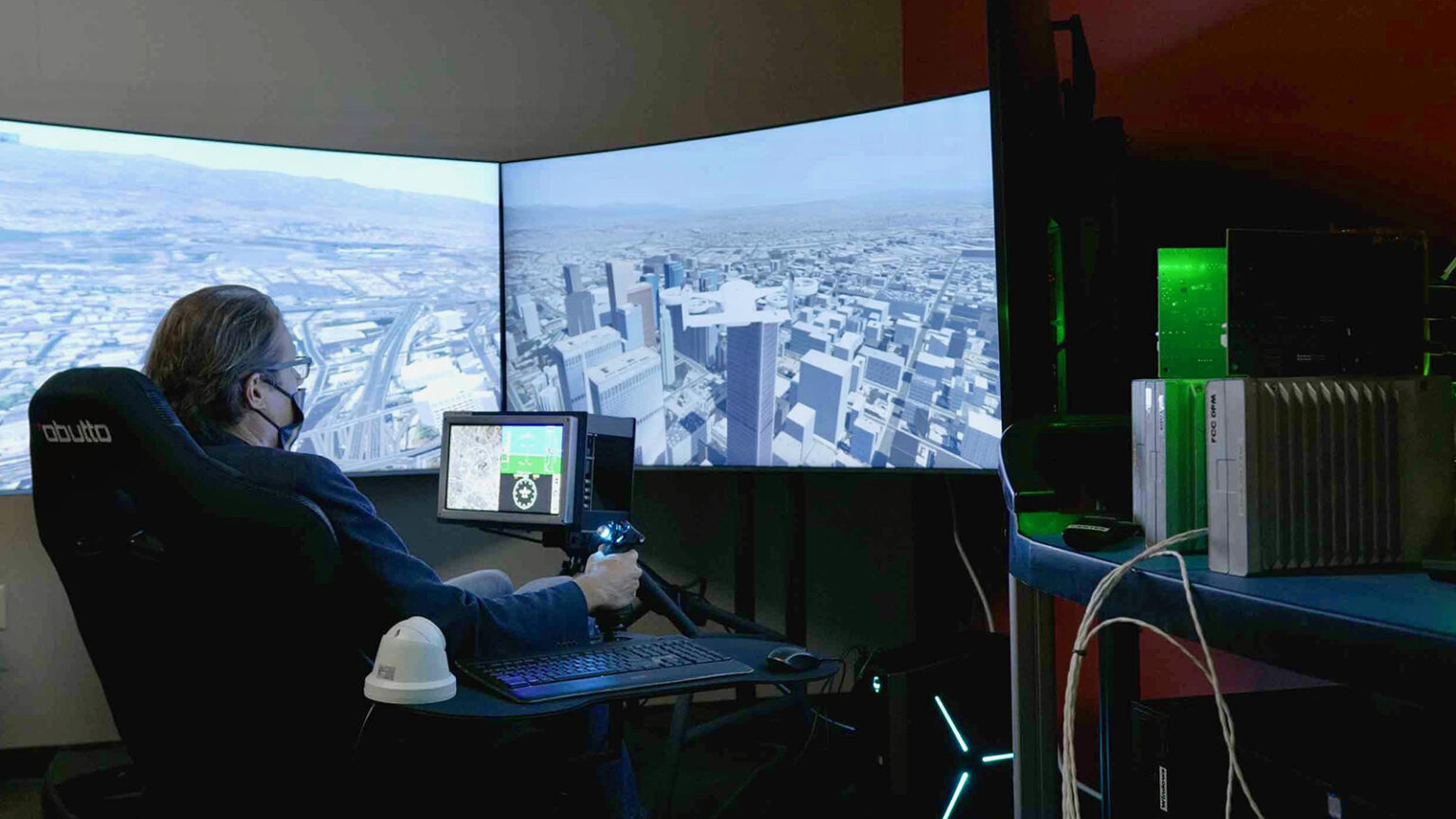 A person sits in a darkened room wearing a headset, operating flight simulation controls in front of multiple large screens displaying a cityscape. Electronic equipment is visible on the right.