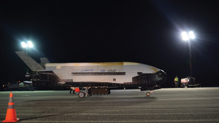 A Boeing X-37B spaceplane sits on an illuminated tarmac at night, showing its USAF markings, with a traffic cone and personnel visible in the scene.