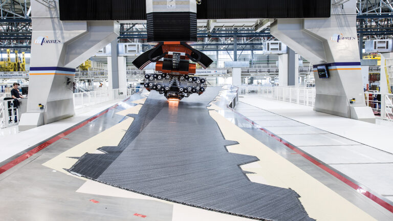 A large industrial weaving machine is laying a carbon fiber material onto a long, flat surface in a modern manufacturing facility.
