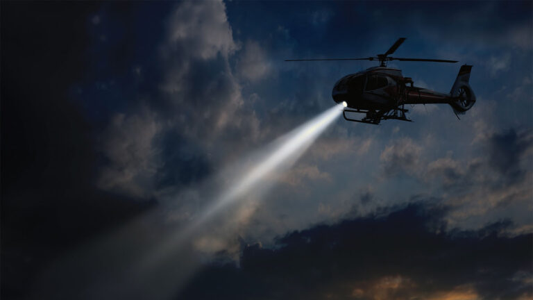 A helicopter with a searchlight hovers in a cloudy night sky.