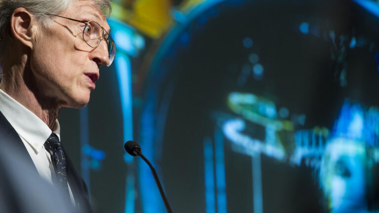 A man with glasses speaks into a microphone, with a blurred, colorful display in the background.