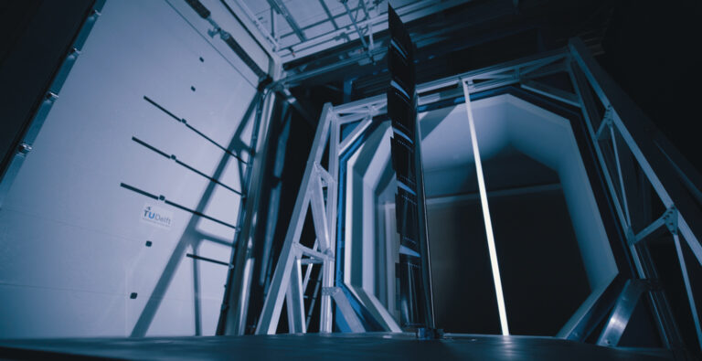 A sleek, modern wind tunnel testing apparatus with metal framing and a vertical panel stands illuminated in a blue-tinted environment.