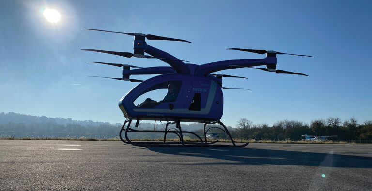 A six-rotor drone aircraft sits on an airstrip under a clear sky, with the sun shining brightly behind it.