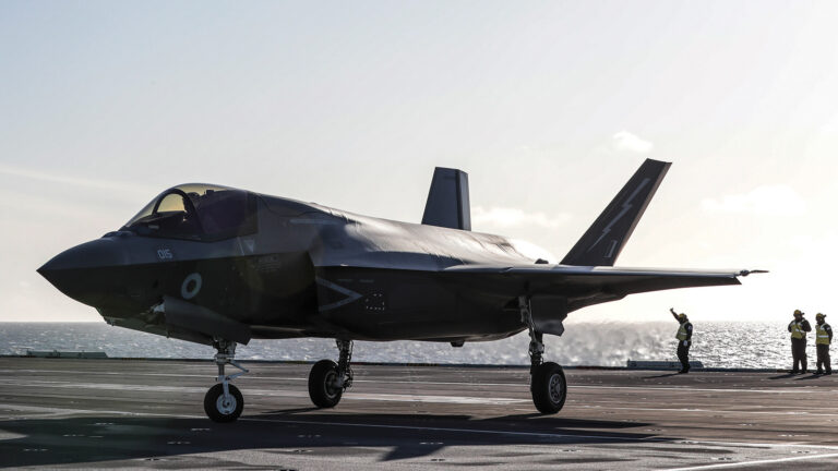 A military fighter jet is taxiing on an aircraft carrier deck with crew members in high-visibility clothing signaling. The ocean is visible in the background under a clear sky.