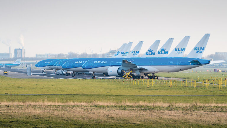 Several KLM Royal Dutch Airlines aircraft are parked in a row on a runway at an airfield, with urban buildings and industrial infrastructure visible in the background.