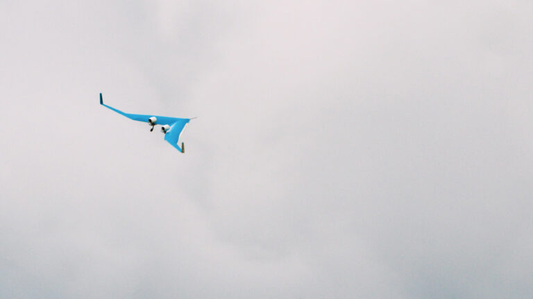 A small, blue drone with distinct wing-like extensions flies against a cloudy sky.