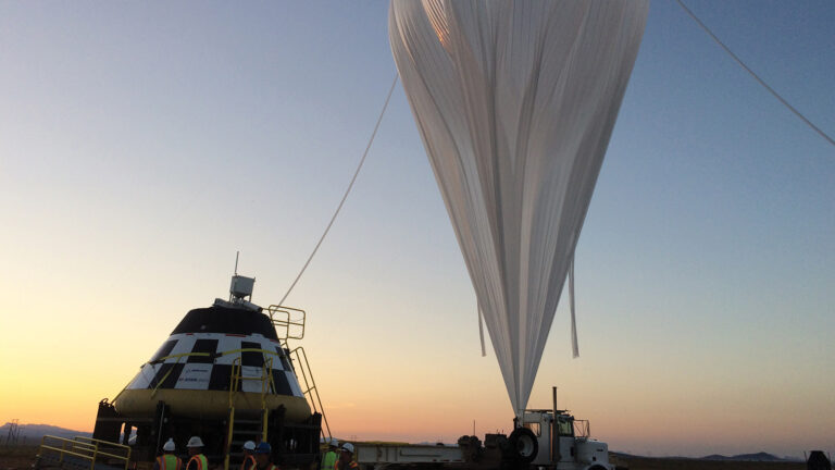A team is preparing a large helium balloon tethered to a capsule-like structure, with the sunset and mountains in the background.