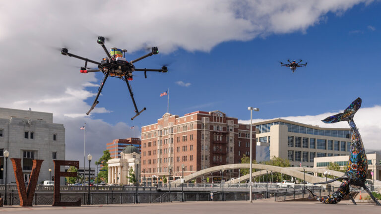 Two drones fly over an urban area with a large sculpture and notable buildings in the background under a partly cloudy sky.