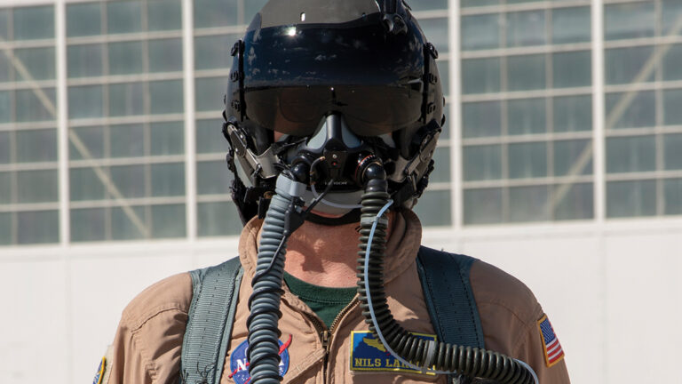 Person in a pilot suit wearing a helmet with a visor and oxygen mask, standing in front of a large grid-windowed building.