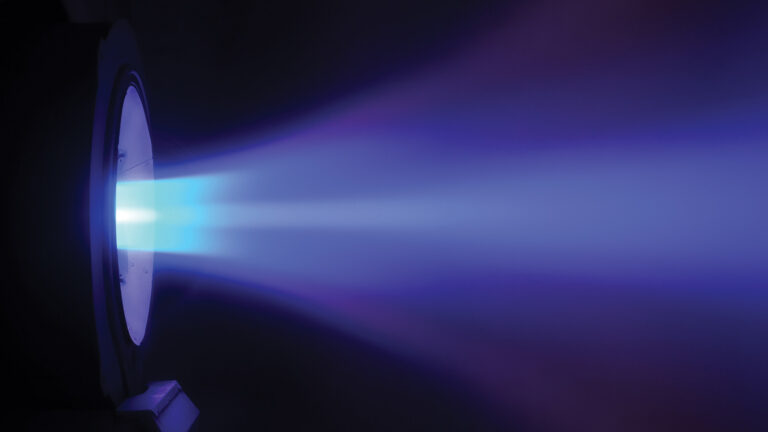 A close-up of a blue and purple ion propulsion system emitting a beam of ionized particles, set against a dark background.