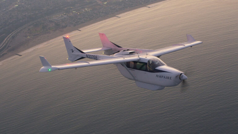 A Ampaire-branded small aircraft is flying over a body of water near a coastline with city infrastructure in the background.