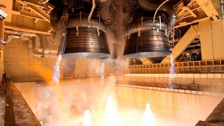 Two rocket engines are seen igniting in a test stand, emitting bright white flames and streams of smoke, with industrial machinery visible in the background.