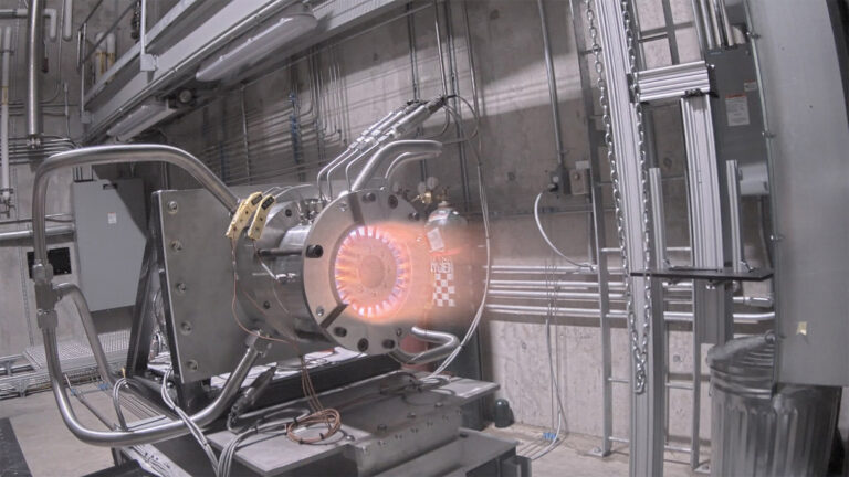 A machine with glowing components and visible wires is housed in an industrial setting with concrete walls and pipes.