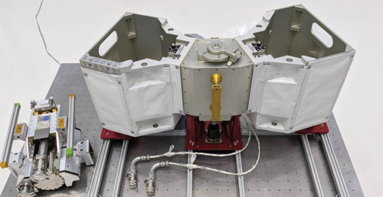 A white and grey mechanical device with various compartments and connectors is placed on a metal surface, alongside a smaller, cylindrical instrument on the left. Both are connected with cables.