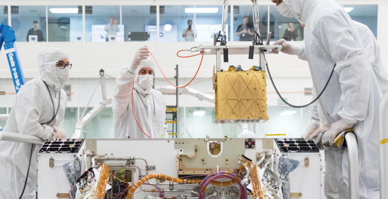 Scientists in cleanroom suits work on assembling or testing a piece of equipment in a laboratory setting, with several onlookers observing from a glass-walled control room above.