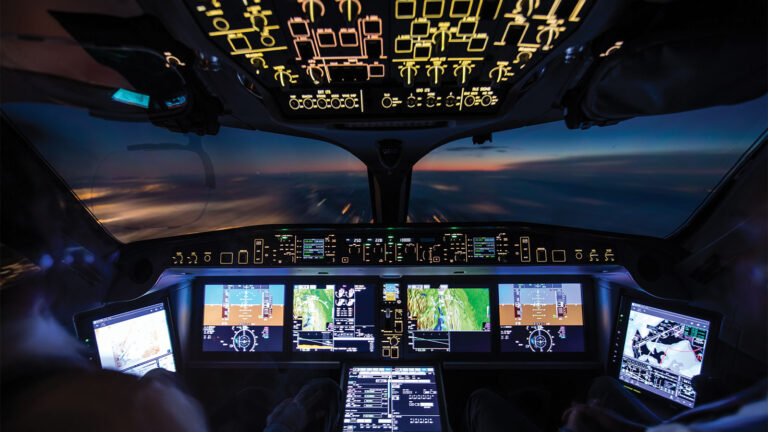 Cockpit view of an aircraft in flight during dusk, with illuminated control panels, screens displaying flight data, and a sunset visible through the windshield.