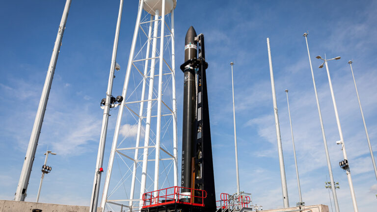 A black rocket stands vertically on a launch pad surrounded by tall structures and a blue sky.