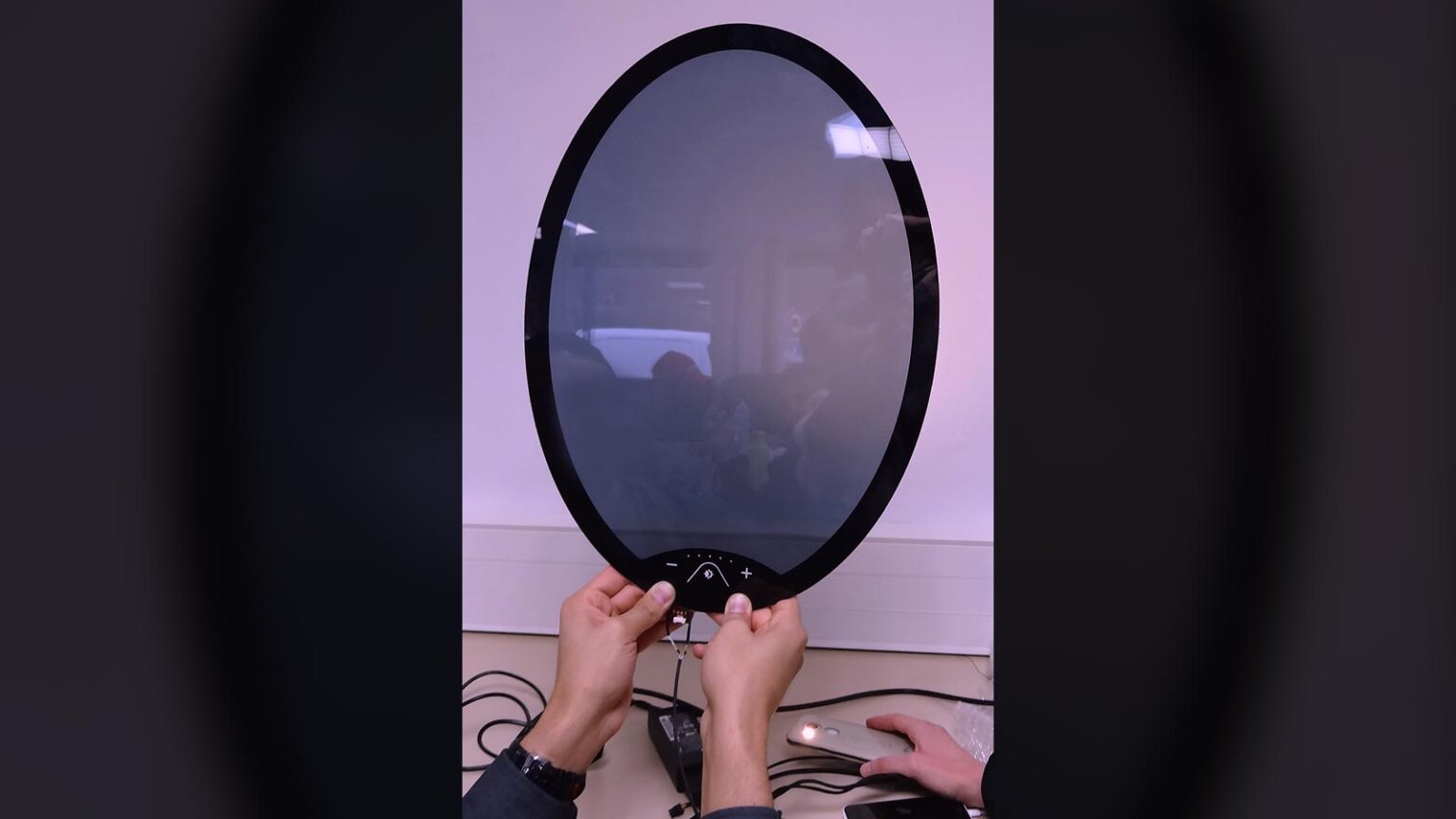 Two hands are seen adjusting a large circular electronic device with a reflective surface, mounted on a table with several wires connected to it.
