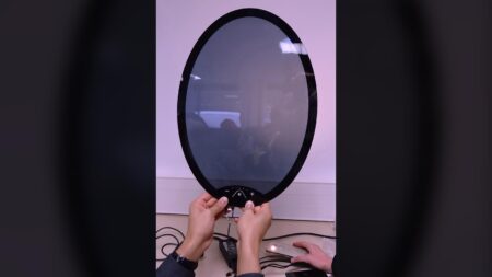 Two hands are seen adjusting a large circular electronic device with a reflective surface, mounted on a table with several wires connected to it.