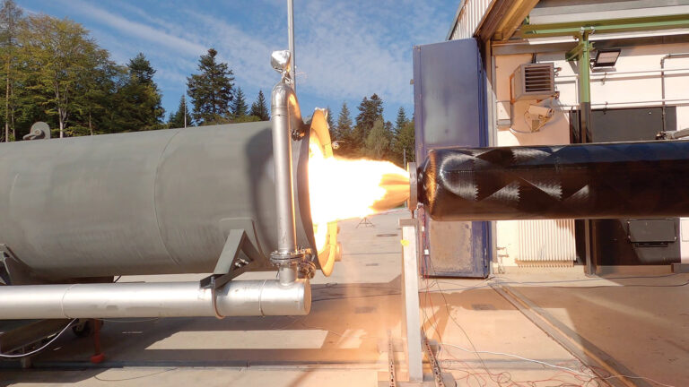 A rocket engine test stand with a lit engine emitting flames is outdoors, surrounded by trees and a clear sky.