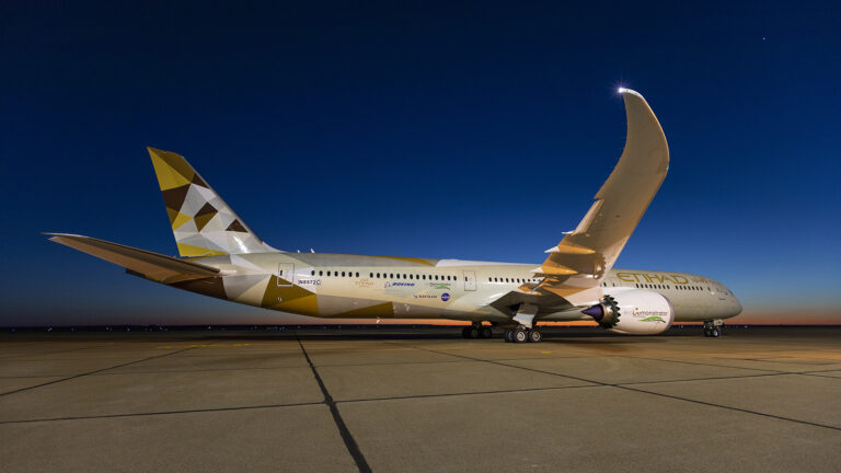 A gold and white Etihad Airways aircraft is parked on a tarmac under a clear evening sky, with its wing prominently curved upwards.