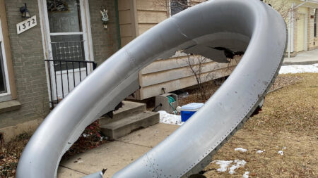 A large, damaged metal ring is seen leaning against the front of a house near the steps. Snow patches are on the ground, and a plastic container sits near the house wall. The house number reads 1372.