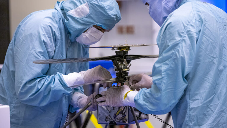 Two people in blue protective suits work on a small helicopter-like device in a cleanroom environment.