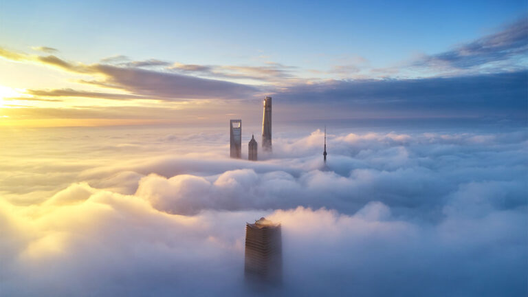 Aerial view of skyscrapers piercing through a thick layer of clouds during sunrise, with a clear sky above and a warm glow on the horizon.