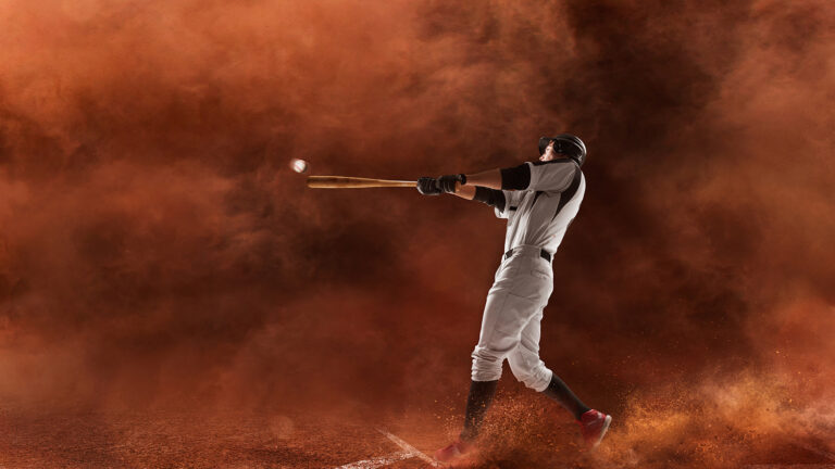 A baseball player in uniform swings a bat at a ball against a background of reddish-orange dust or smoke.