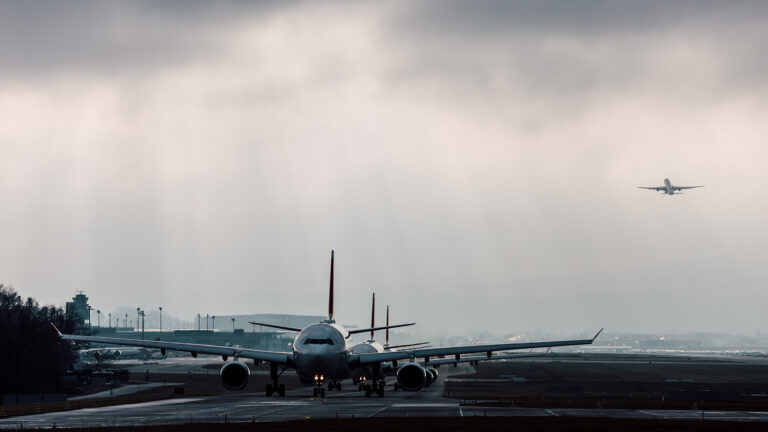 Two airplanes taxiing on the runway with another plane taking off in the background under a cloudy sky.