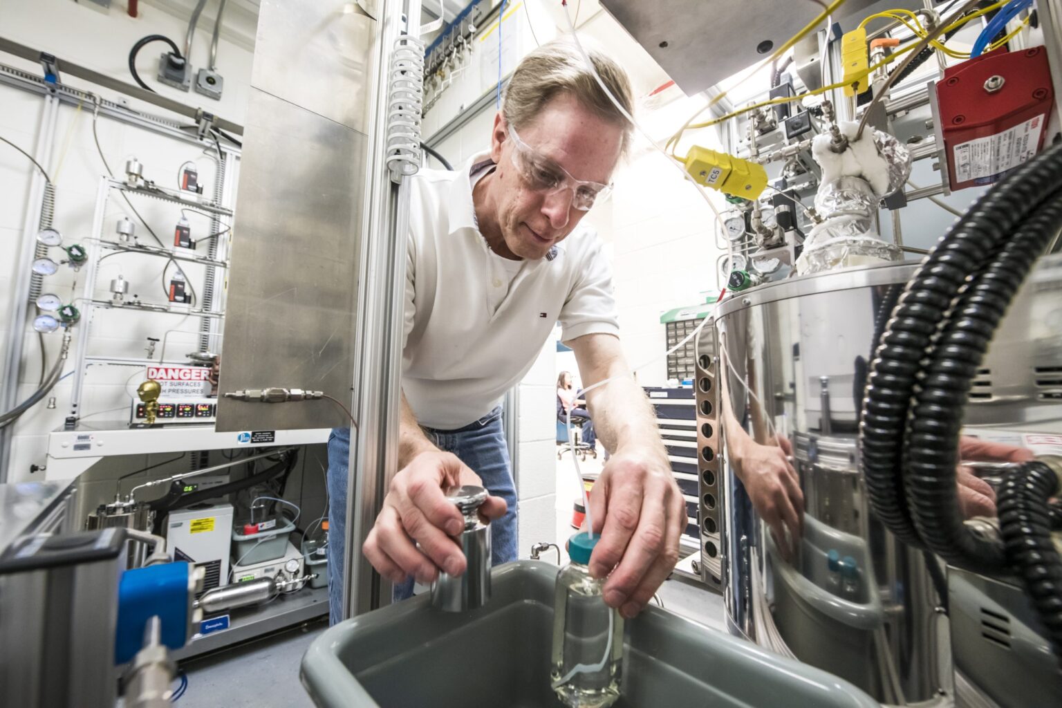 A man wearing safety glasses and a white polo shirt works with laboratory equipment, handling glassware near a large machine in an industrial laboratory setting.