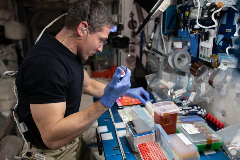 An individual wearing gloves and a headset works with scientific equipment and containers in a cluttered workspace, likely conducting an experiment.