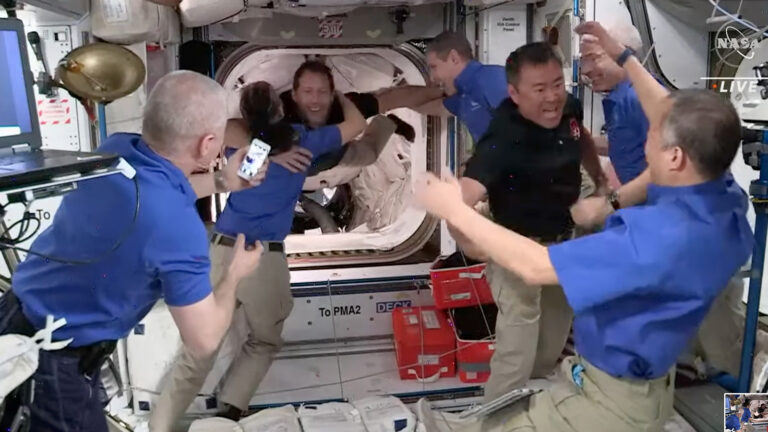 Astronauts inside the International Space Station greet and embrace arriving crew members in a zero-gravity environment, with one person holding a phone.