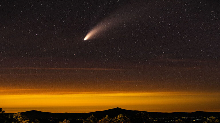A bright comet streaks across a night sky filled with stars above a silhouetted mountain range, with an orange glow near the horizon.