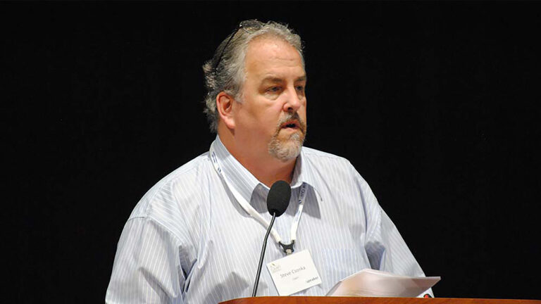 A person with gray hair and a beard stands behind a podium, speaking into a microphone, holding papers. They are wearing a light blue striped shirt and a badge around their neck.