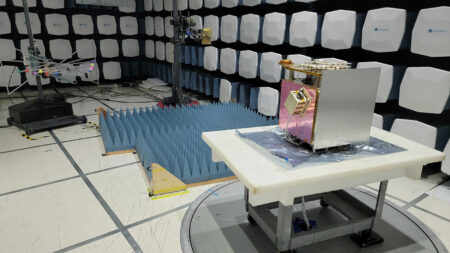 A small satellite undergoing testing in an anechoic chamber. The room is lined with sound-absorbing panels, and the satellite is placed on a turntable on a foam pad.