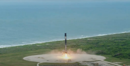 A rocket booster is landing vertically on a concrete pad near the coastline, emitting flames from its base during touchdown.