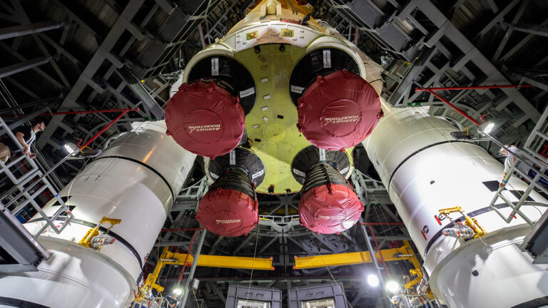 A close-up view of the base of a large rocket, showing red protective covers on the rocket engines and large cylindrical fuel tanks around it, inside a facility with various equipment and scaffolding.