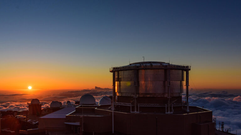 A large observatory with domes is silhouetted against a colorful sunset sky, with clouds below the horizon.